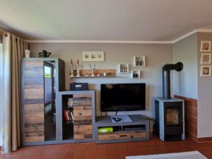 Offene Küche. Wohnzimmer mit Schlafcouch für zwei Personen und Couchtisch, Wohnzimmerwand, Esstisch mit Sitzbank und drei Stühlen, Kaminofen. Zur Unterhaltung stehen ein Flachbild-TV mit Sat-Empfang, ein DVD-Player und ein Radio zur Verfügung.