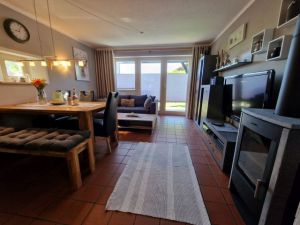 Offene Küche. Wohnzimmer mit Schlafcouch für zwei Personen und Couchtisch, Wohnzimmerwand, Esstisch mit Sitzbank und drei Stühlen, Kaminofen. Zur Unterhaltung stehen ein Flachbild-TV mit Sat-Empfang, ein DVD-Player und ein Radio zur Verfügung.